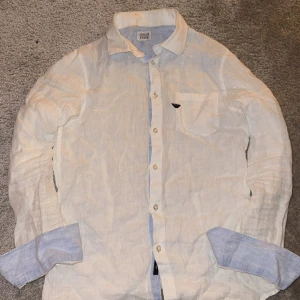 Vit linneskjorta från Armani Jeans - Vit skjorta från Armani Jeans i mjukt linne med klassisk krage och knappar framtill. Ljusblå detaljer på insidan av manschetterna och knappslån. Bröstficka med svart Armani-logga. Perfekt för dig som gillar stilrena och fräscha plagg.