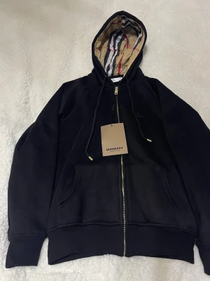 Svart burberry zip  - Svart hoodie från Burberry med klassisk rutmönstrad huva i beige, rött och svart. Tröjan har guldfärgad dragkedja, snörning vid huvan och broderad Burberry-logga på bröstet. Tillverkad i mjuk bomull och har praktiska fickor framtill