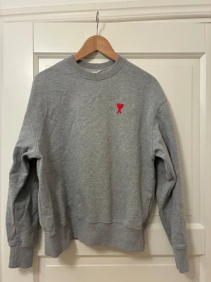 Grå sweatshirt från Ami Paris - Grå crewneck sweatshirt från Ami Paris med röd broderad logga med hjärta och A på bröstet. Klassisk passform med ribbade muddar vid ärmslut, hals och nederkant. Mycket bra skick!