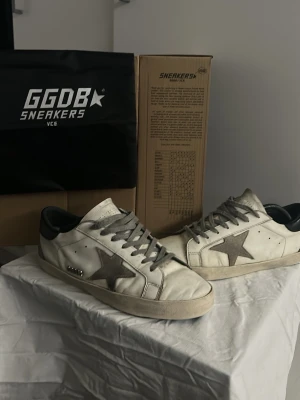 GGDB vita sneakers med grå stjärna - Säljer ett par GGDB sneakers i vitt skinn med grå snörning och en grå stjärna på sidan. Skorna har svart hälparti och platt sula. Klassisk låg modell med rund tå och coola metall-detaljer vid sidan. Perfekta för dig som gillar streetstyle och vill ha något unikt.