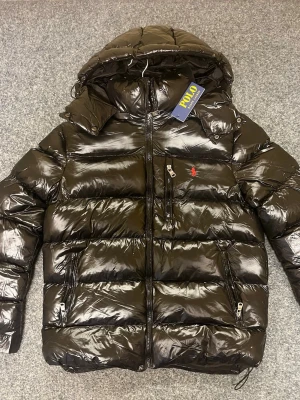 Svart pufferjacka från Polo Ralph Lauren - Säljer en svart, glansig pufferjacka från Polo Ralph Lauren med huva och flera dragkedjeförsedda fickor. Jackan har röd Polo-logga på bröstet och insidan, samt justerbara detaljer vid huvan och nederkant. Perfekt för kalla dagar och riktigt snygg streetstil.