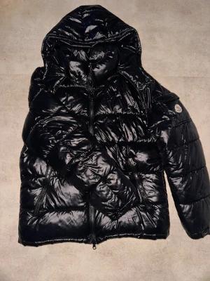 Svart Moncler pufferjacka med huva - Säljer en svart, glansig pufferjacka från Moncler i storlek M. Jackan har huva, två dragkedjefickor fram och ikonisk Moncler-logga på ärmen. Quiltad design och hel dragkedja framtill. Perfekt för kalla dagar och riktigt snygg streetstil.