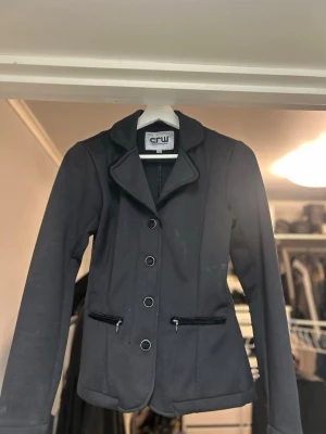 Svart ridkavaj från CRW, stl 36 - Snygg svart ridkavaj från CRW med klassisk krage och fyra velour/silverfärgade knappar framtill. Jackan har två fickor med dragkedja och dekorativa knappar vid ärmslut. Materialet är mjukt och följsamt, perfekt för ridning. Passformen är normal och ärmarna är normallånga. Kavajen är använd mycket och defekter finns se bild 4 & 5 då det skavits med ridvästen, men i helhet en väldigt fin kavaj. Stl 36