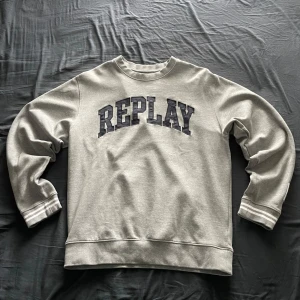 Grå sweatshirt från Replay - Säljer en grå sweatshirt från Replay med tryckt logga på bröstet och patch på ärmen. Tröjan har rund halsringning och ribbade muddar vid ärmslut och nederkant. Perfekt för en avslappnad stil.