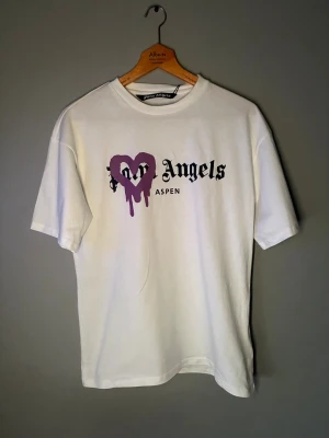Palm Angels Aspen vit t-shirt - Palm Angels Aspen t-shirt i vit bomull med tryck framtill. Har ett stort lila hjärta med drip-effekt och svart text i gotisk stil. Klassisk rund hals och korta ärmar. Perfekt statement-plagg för dig som gillar streetwear och snygga prints.