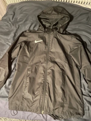 Svart Nike Storm-Fit vindjacka L - Svart vindjacka från Nike med Storm-Fit teknologi, perfekt för blåsiga och regniga dagar. Jackan har huva, hel dragkedja framtill och två sidofickor. Swoosh-logga på bröstet och justerbar nederkant med snörning. Lätt och sportig design.