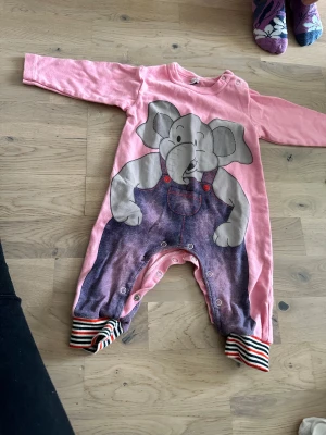 Rosa byxdress med Dumbo-tryck - Rosa sparkdräkt med en elefantillustration som bär jeansoveraller. Sparkdräkten har långa ärmar och randiga muddar vid bensluten. Den har tryckknappar längs framsidan och benen för enkel på- och avklädning. 3-12 månader Söt rosa byxdress med lång ärm och tryck av Dumbo i lila hängselbyxor. Knäppning med tryckknappar längs benen och axeln för enkel påklädning. Muddar i rött, svart och vitt randigt mönster vid bensluten. Materialet är mjukt bomullstyg.