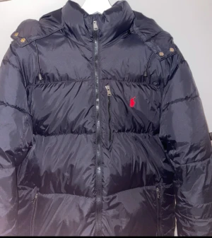 Svart pufferjacka från Ralph Lauren - Svart pufferjacka från Ralph Lauren med röd broderad logga på bröstet. Jackan har huva, dragkedja framtill, flera fickor med dragkedja och quiltad design. Perfekt för kalla dagar och har en klassisk, sportig look. 