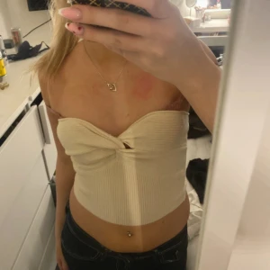 Beige ribbad bandeau tubtopp - Säljer en beige ribbad bandeau tubtopp med twistad detalj framtill. Toppen är ärmlös och croppad, perfekt för en trendig look. Sitter tight och har en snygg passform som framhäver axlarna.