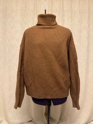 Brun polotröja från H&M, storlek M - Mysig brun polotröja från H&M i storlek M. Tröjan har en hög krage, långärmade och är stickad i ett mjukt material. Passformen är loose och den har ribbade muddar vid ärmslut och nederkant. Perfekt för kyliga dagar.