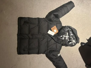 Svart pufferjacka från Burberry - Svart pufferjacka från Burberry London England med stor logopatch på ärmen. Jackan har huva med rutigt foder i svart, vitt och lite rött. Flera fickor med dragkedja och knappstängning framtill. Perfekt för kalla dagar och riktigt snygg streetstil.