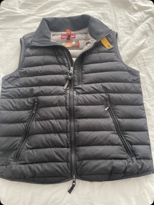 Svart dunväst från Parajumpers - Snygg svart dunväst från Parajumpers med quiltad design och dragkedja framtill. Västen har två praktiska fickor med dragkedja och en gul detalj vid kragen. Insidan är ljusgrå och märket syns tydligt på både insidan och utsidan. Perfekt för lager-på-lager under höst och vinter.