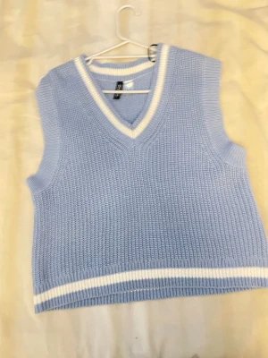 Ljusblå stickad väst med v-ringning - Supersnygg ljusblå stickad väst från Gina Tricot med vit kant vid v-ringningen och nederkanten. Västen har en loose passform och är perfekt att styla över en skjorta eller t-shirt. Mjukt material och klassisk design med sportig touch.