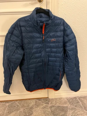 Mörkblå quiltad pufferjacka från Five - Säljer en mörkblå pufferjacka från Five i storlek L. Jackan har quiltad design, orange detaljer vid dragkedja och nederkant, samt hög krage. Perfekt för kyliga dagar och riktigt snygg med sin sportiga look.