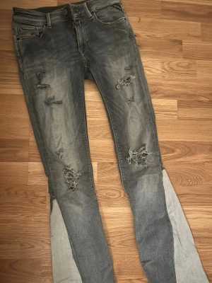 Replay jeans med slitningar - Säljer ett par Replay jeans i blå tvätt med slitna detaljer. Jeansen har klassisk femficksmodell, låg midja och är tillverkade i stretchig bomullsmix för skön passform. Perfekta för dig som gillar en edgy och avslappnad stil.