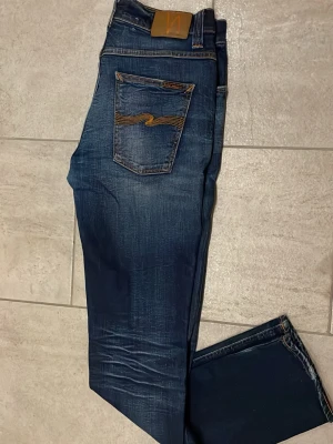Blå jeans från Nudie Jeans - Säljer ett par snygga blå jeans från Nudie Jeans i storlek 33/34. Jeansen har klassisk femficksdesign, orangea sömmar och coola broderade bakfickor. Modellen är rak med smal passform och tillverkad i slitstarkt bomullsmaterial.
