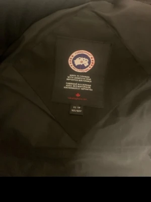 Svart dunväst från Canada Goose XS - Säljer en svart dunväst från Canada Goose i storlek XS passar i S också. Västen har hög krage, två stora fickor framtill och stängs med både dragkedja och knappar. Klassisk Canada Goose-logga på bröstet och riktigt varm dunfyllning.