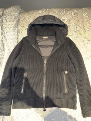 Svart Moncler pufferjacka med stickade ärmar - Svart pufferjacka från Moncler med stickade ärmar och huva. Jackan har två fickor med dragkedja och ribbad kant vid ärmslut och nederkant. Framsidan har en tydlig Moncler-logga och jackan är quiltad på ryggen och huvan. Perfekt för kyliga dagar.