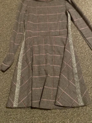 Rutig klänning med glitter från Yigga - Supersnygg rutig klänning från Yigga i grått med rosa och röda linjer. Klänningen har lång ärm och glittriga silverdetaljer längs kjolen. Materialet är mjukt och stretchigt, perfekt för dig som gillar att sticka ut med coola detaljer.