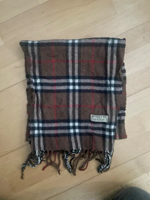 Brun rutig Burberry halsduk i ull - Säljer en brun halsduk från Burberry med klassiskt rutigt mönster i svart, vitt och rött. Halsduken är tillverkad i 100% lammull och har fransar längs nederkanten. Perfekt för dig som gillar tidlös design och vill ha något som sticker ut. Perfekt för vintern. Längd:135cm bredd 35cm