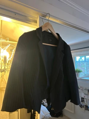 Svart randig kavaj - Snygg svart kavaj med diskreta vita ränder. Klassisk modell med krage och knäppning framtill. Passar mig som vanligtvis har 34 i storlek.