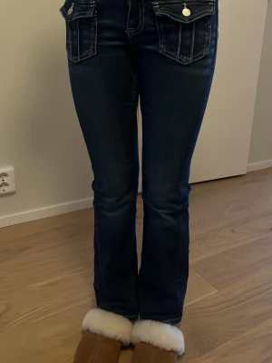Mörkblå bootcut jeans med fickdetaljer - Snygga mörkblå jeans med bootcut-snitt och markerade fickor fram med lock och silverknappar. Jeansen har klassisk femficksdesign och kontrastsömmar som ger en cool look. Perfekta för dig som gillar jeans med lite extra detaljer och stil.