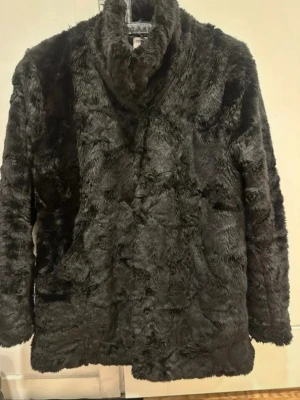 Svart pälsjacka från Tiger of Sweden stl S - Mjuk och fluffig svart pälsjacka från Tiger of Sweden med hög krage och långärmad design. Jackan har en clean look utan synliga knappar eller dragkedja och är tillverkad i syntetmaterial. Perfekt för dig som vill ha en stilren och cozy vibe.