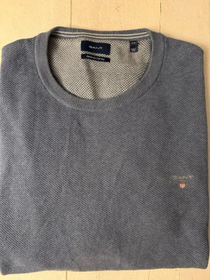 Grå bomullströja från GANT - Snygg grå tröja från GANT i premium bomull med rund hals och diskret logga broderad på bröstet. Tröjan har en klassisk, normal passform och är långärmad. Perfekt för dig som gillar stilrena och tidlösa plagg.