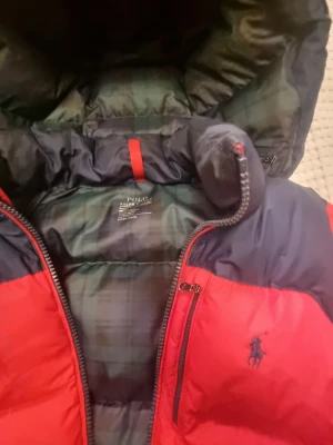 Röd och marinblå jacka Polo Ralph Lauren - Jacka från Polo Ralph Lauren i rött och marinblått. Jackan har huva, dragkedja framtill och sidofickor med dragkedja. Insidan har rutigt foder och klassisk Polo-logga på bröstet. Knappt märkbar fläck bredvid vänstra fickan samt under ena ärmen. Se bilder. Därav priset.