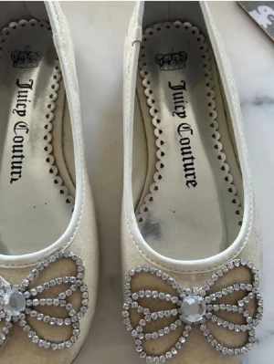 Glittriga ballerinaskor Juicy Couture - Säljer ett par vita ballerinaskor från Juicy Couture med glittrande rosettdetaljer framtill i strass. Skorna har rund tå och är tillverkade i ett skimrande material som ger en lyxig känsla. Perfekta för dig som vill sticka ut lite extra.