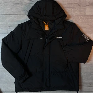 Svart pufferjacka från Timberland - Säljer en svart pufferjacka från Timberland med huva och dragkedja. Jackan har långa ärmar och en broderad Timberland-logga på bröstet och ärmen. Perfekt för kyliga dagar.