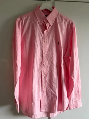 Rosa skjorta från Ralph Lauren - Klassisk rosa skjorta från Ralph Lauren med button-down krage och broderad logga på bröstet. Skjortan har långa ärmar, vita knappar och är gjord i mjuk bomull. Perfekt för en clean och stilren look. Den är lite smutsig vid vänster armhåla, därav priset. 