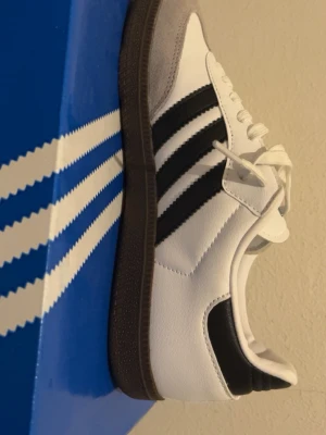 Adidas Samba vita och svarta sneakers - Adidas Samba sneakers i klassisk vit och svart färg med bruna sulor. Skorna har tre svarta ränder på sidan, snörning och detaljer i grå mocka vid tån. Ovandelen är i skinn och textil, med rund tå och platt sula. Perfekt för dig som gillar retrostil och ikoniska modeller.