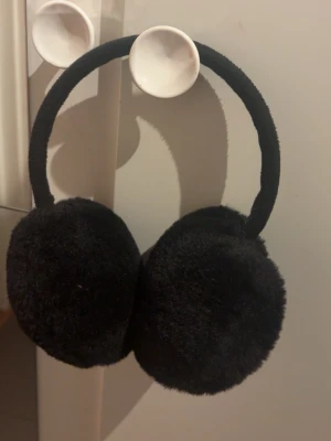 Svarta öronmuffar - Säljer svarta öronmuffar med rundad design och mjukt material. Muffarna har en enkel och stilren look som passar dig som gillar att sticka ut med fluffiga detaljer. Perfekt för att addera lite extra värme och stil till din outfit.