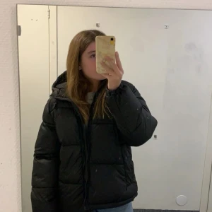 Svart pufferjacka med huva - Säljer en svart pufferjacka med huva och dragkedja. Jackan är vadderad och har en oversized passform. Perfekt för kalla dagar när du vill hålla dig varm och ändå se snygg ut. Den har några sömmar som gått upp i ryggen vilket knappt syns. 