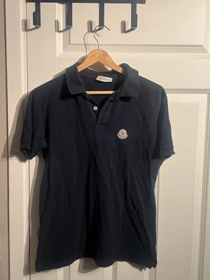 Mörkblå Moncler pikétröja - Säljer en stilren mörkblå pikétröja från Moncler med klassisk krage och två knappar framtill. Tröjan har korta ärmar och ett broderat Moncler-märke på bröstet. Perfekt för dig som gillar enkel och snygg design.