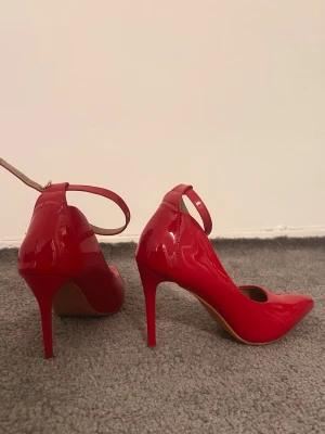 Röda pumps med smal klack - Säljer ett par snygga röda pumps med smal hög klack och spetsig tå. Skorna har en glansig finish och ett elegant ankelband med spänne. Perfekta för dig som vill sticka ut med färg och stil.