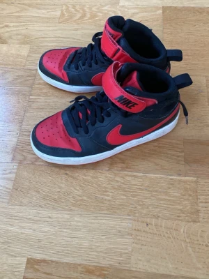 Svarta och röda Nike high sneakers - Säljer ett par svarta och röda Nike sneakers med högt skaft och klassisk swoosh på sidan. Skorna har snörning, kardborreband upptill och vit sula. Perfekta för dig som gillar streetwear och vill sticka ut med en ikonisk färgkombo.