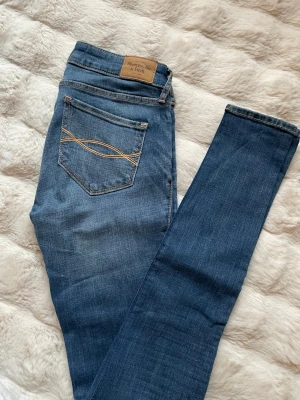 Abercrombie & Fitch Super Skinny Jeans - Säljer ett par blå super skinny jeans från Abercrombie & Fitch med klassisk femficksdesign och snygga kontrastsömmar. Modellen har låg midja och smal passform hela vägen ner. Materialet är mjukt jeans med stretch för extra komfort.