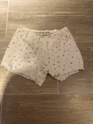 Blommiga ribbade shorts XS - Söta vita shorts från C i storlek XS med små blå blommor och diskreta ränder. Ribbad struktur och volangkant nedtill ger en gullig vibe. Perfekta för varma dagar eller som mysplagg hemma. Gjorda i mjuk bomull med lite stretch.