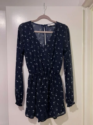 Mörkblå blommig byxdress med lång ärm - Säljer en mörkblå byxdress från Hollister med små vita blommor, omlott framtill och resår i midjan. Långa ärmar med rynkade muddar och lätt, luftig polyester. Perfekt för dig som gillar en chill och snygg stil.