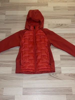 Röd pufferjacka från Everest - Säljer en röd pufferjacka från Everest med huva och hel dragkedja. Jackan har quiltad framsida och baksida, samt släta ärmar. Perfekt för dig som gillar sportig stil och vill ha något som sticker ut i höst och vinter.