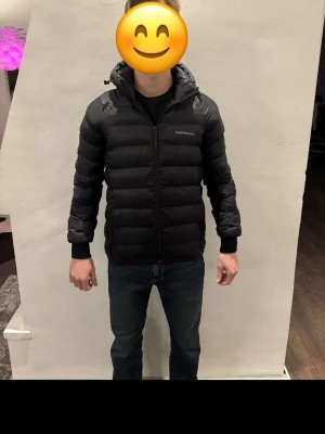 Svart Peak Performance dunjacka - Svart dunjacka från Peak Performance med huva och dragkedja framtill. Jackan har en quiltad design och ribbade muddar vid ärmslut. Perfekt för kalla dagar och har en clean look som funkar till jeans och sneakers. Modellen är 175 cm, vikt 68 kg
