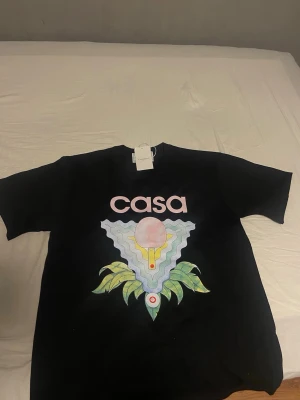 Casablanca tröja - Svart t-shirt från Casablanca med coolt grafiskt tryck framtill och texten 'casa' i rosa. Trycket har vågiga former, pastellfärger och gröna blad. T-shirten har klassisk rund hals och korta ärmar. Perfekt för dig som gillar streetwear och snygga prints.
