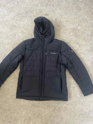 Svart pufferjacka från Peak Performance - Svart pufferjacka från Peak Performance med huva och dragkedja framtill. Jackan har quiltad design och en bröstficka med zip. Perfekt för kalla dagar och har en clean, sportig look som funkar till många stilar.