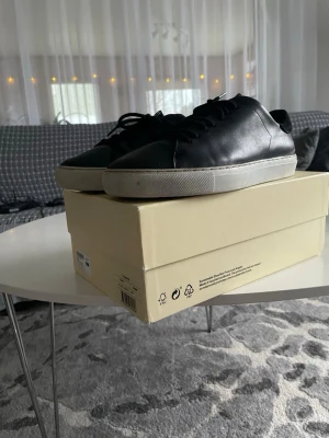 Svarta Axel Arigato Clean 90 sneakers - Säljer ett par svarta Axel Arigato Clean 90 sneakers i skinn med klassisk vit platt sula. Skorna har minimalistisk design, rund tå och svarta snören. Perfekta för dig som gillar stilrena och tidlösa sneakers. Box och originalförpackning medföljer.