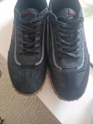 Svarta sneakers från Hugo med röd logga - Säljer ett par svarta sneakers från Hugo med röd logga på sidan och plösen. Skorna har snörning, detaljer i grått och en brun sula. Materialet är en mix av mocka och syntet, vilket ger en sportig och stilren look.