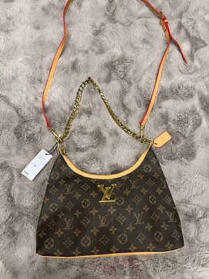 Louis Vuitton monogram axelväska - Snygg axelväska från Louis Vuitton med klassiskt monogrammönster i brunt och beige. Väskan har guldfärgad kedja, justerbar axelrem med röda detaljer och LV-logga framtill. Tillverkad i skinn med exklusiv känsla och fina detaljer.