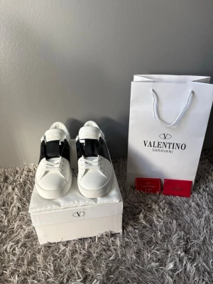 Valentino Garavani vita sneakers - Säljer ett par snygga Valentino Garavani sneakers i vitt med svart detalj över sidan. Skorna har rund tå, platt sula och klassisk snörning. Tillverkade i skinn och kommer med originalförpackning och certifikat. Perfekt för dig som gillar lyxiga och stilrena sneakers. Helt oanvända.