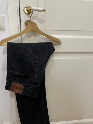 Svarta jeans från Tommy Hilfiger - Säljer ett par svarta jeans från Tommy Hilfiger med klassisk femficksdesign och snygg läderpatch bak. Jeansen har rak passform och är tillverkade i slitstarkt denim-material. Perfekt för dig som gillar stilrena och tidlösa plagg.